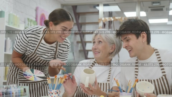 A cheerful elderly woman, along with a young girl and a guy wearing aprons, are engaged in ceramic dishware painting during a pottery class. This is a creative hobby and an intergenerational communication concept. Стоковое видео, видеограф Яков Филимонов / Фотобанк Лори