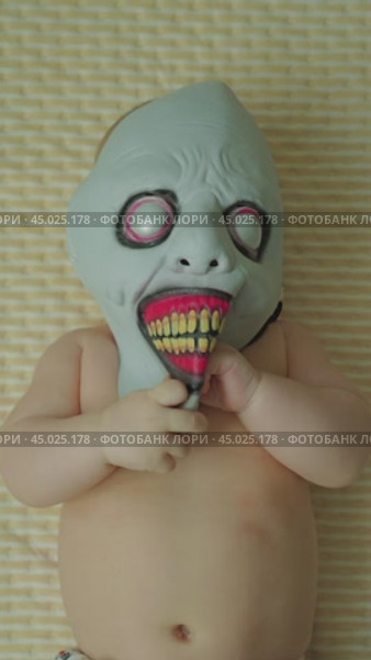 A child wearing a creepy monster mask with red gums and big teeth lies on his back. Small hands touch the mask and crumple it. The baby's shirt torso is visible. Creepy baby. Стоковое видео, видеограф Воловодов Владимир / Фотобанк Лори