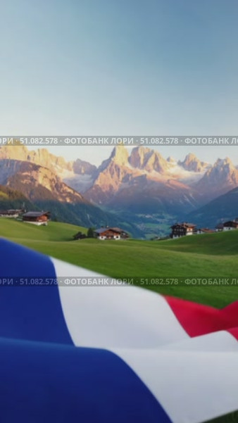 A close-up of the French flag waving in the foreground with a scenic mountain landscape in the background. The sky is clear and blue, enhancing the vibrant colors of the flag. Стоковое AI-видео, аниматор Nart / Фотобанк Лори