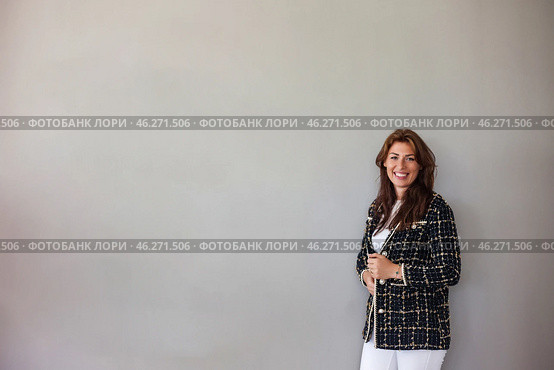 A confident woman with long, flowing hair stands beside a smooth gray wall, dressed in an elegant blazer and white pants. Her cheerful smile adds warmth to the minimalist atmosphere of the room. Стоковое фото, фотограф Aleksandr Golubev / Фотобанк Лори