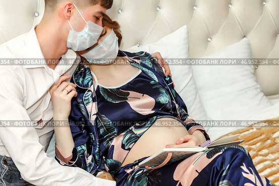 A couple sits closely together on a plush bed, both wearing masks. The woman, visibly pregnant, reads a book while resting against her partner, creating an intimate atmosphere. Стоковое фото, фотограф Aleksandr Golubev / Фотобанк Лори