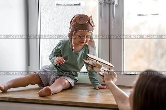 A cute 2-year-old boy in a cap is playing on the windowsill with a wooden airplane. Children's wooden toys for child development. Стоковое фото, фотограф Photojuli / Фотобанк Лори