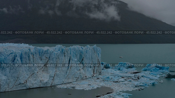 A fragment of a melting glacier. A wall of cracked blue ice rises above a turquoise glacial lake. Melted ice floes float in the water. Mountains in fog and clouds. Perito Moreno. El Calafate.Argentina. Стоковое фото, фотограф Вера Ивановна Кубарева / Фотобанк Лори