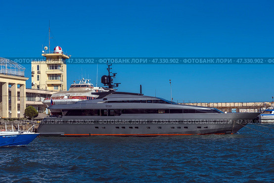 A fragment of the seaport in Yalta. In the center of the frame, an elegant and modern yacht called Orion is moored, painted dark gray with an orange line along the waterline. Nearby are boats and other vessels, and in the background are visible buildings of the port infrastructure, including the control tower with a characteristic dome. The sea is bright and rich blue, light ripples on the water indicate a fresh breeze. On the concrete wall behind is the inscription WELCOME, which emphasizes the tourist character of the place.The photo conveys the atmosphere of active marine life and the modern Yalta coast. (2021 год). Редакционное фото, фотограф Анна Кривицкая / Фотобанк Лори