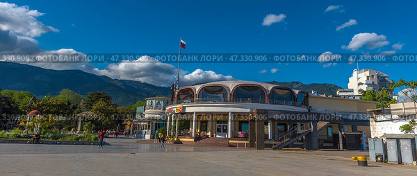 A fragment of the Yalta embankment, one of the most popular resort towns in Crimea. In the foreground is a modern building with a round façade and stained-glass windows, where cafes and restaurants are located. On the left is part of the pedestrian zone with passers-by, vacationers and strollers along the shore. In the background, the green mountains of the Crimean ridge rise, and the sky is decorated with living clouds and a flying bird. The Russian flag on the mast emphasizes the formality and maritime character of the place.The decor conveys an atmosphere of relaxation, resort activity and harmony with the natural landscape. (2021 год). Редакционное фото, фотограф Анна Кривицкая / Фотобанк Лори