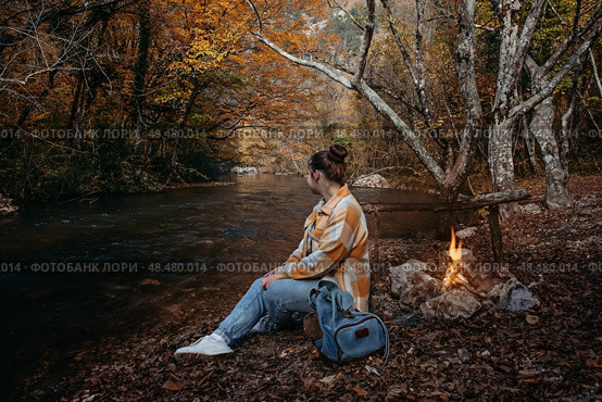 A girl in a plaid shirt sits on a log on the river bank by the fire. Стоковое фото, фотограф София Тюленева / Фотобанк Лори