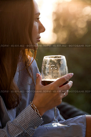 A glass of red alcoholic drink or wine in a woman's hand. A woman is resting on the veranda of a country house. The concept of relaxation, party or celebration. Стоковое фото, фотограф Photojuli / Фотобанк Лори