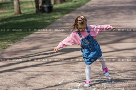 A happy girl in pink clothes draws classics with chalk on the asphalt on the street. Portrait of a little girl playing and jumping hopscotch on a sunny summer day. Creative development of children. Стоковое фото, фотограф Photojuli / Фотобанк Лори