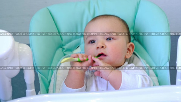A hungry baby is gnawing on a plastic spoon at the table on a high chair. Teething, whims, itchy gums, introduction of complementary foods. Стоковое видео, видеограф Ольга Симонова / Фотобанк Лори