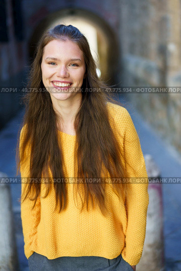 A joyful young woman with long, flowing hair, dressed in a vibrant and bright yellow sweater, stands in a picturesque urban alley, radiating positivity, warmth, and happiness like sunshine. A cheerful woman in a yellow sweater posed in an urban setting with vibrant city colors. Стоковое фото, агентство Ingram Publishing / Фотобанк Лори