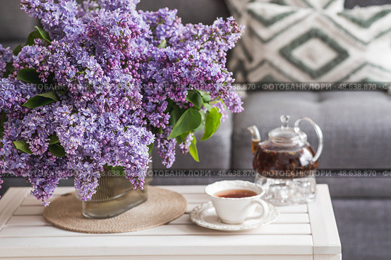 A large purple bouquet of lilacs and a cup of tea and a teapot on the table. In the background is the interior of a modern living room. The concept of home comfort . Стоковое фото, фотограф Photojuli / Фотобанк Лори