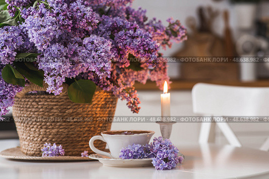 A large purple bouquet of lilacs in a wicker basket, a cup of tea and candles on the table. In the background is the interior of a modern white kitchen. The concept of home comfort. Стоковое фото, фотограф Photojuli / Фотобанк Лори