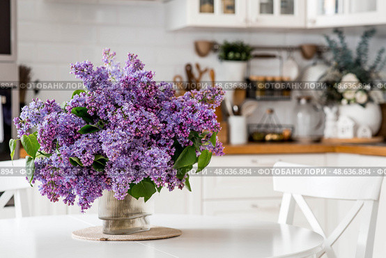 A large purple bouquet of lilacs on the table. In the background is the interior of a modern white Scandinavian-style kitchen. The concept of home comfort and celebration. Стоковое фото, фотограф Photojuli / Фотобанк Лори