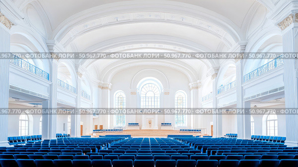 A majestic architectural interior of a bright, pristine white assembly hall, featuring grand vaulted ceilings, elegant arches, and rows of empty blue seating, prepared for a formal event or solemn ga. Стоковое AI-изображение, AI-художник Данила Большаков / Фотобанк Лори