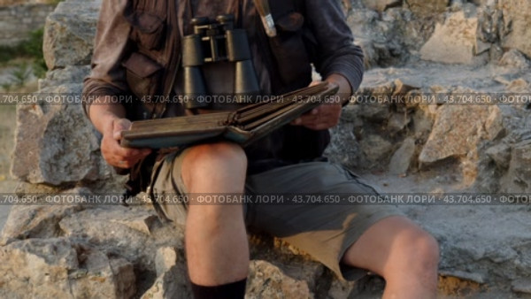 A man dressed as a researcher and with binoculars sits on the rocks and studies an old book. Стоковое видео, видеограф Константин Скуридин / Фотобанк Лори