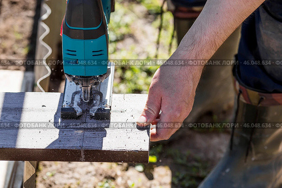 A man holds a battery-powered electric saw and builds new beds in a greenhouse made of wood-polymer composite. Macro photography. Side view. Preparing for the spring garden season. Стоковое фото, фотограф Photojuli / Фотобанк Лори