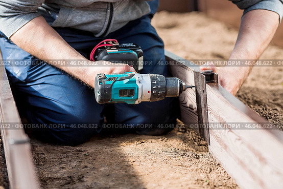 A man holds a battery-powered screwdriver and collects new beds from WPC into a greenhouse made of polyethylene, polycarbonate. Macro photography. Side view. Preparing for the spring garden season. Стоковое фото, фотограф Photojuli / Фотобанк Лори