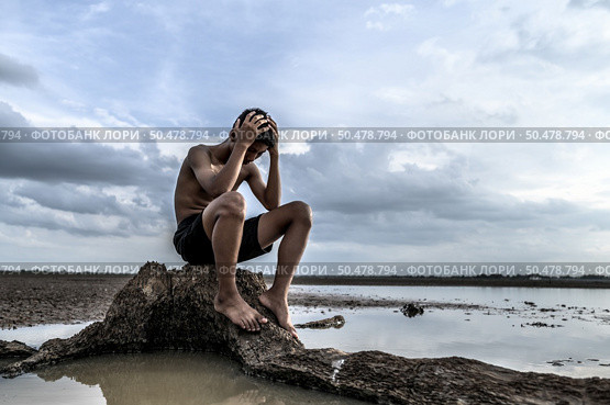 A man sat bent his knees and put his hands on his head, on the base of the tree and surrounded by water. Стоковое фото, агентство Ingram Publishing / Фотобанк Лори