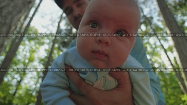 A man tilts a infant in a blue onesie towards the camera against a sunlit forest background, a close-up portrait with brightly coloured eyes and ruddy cheeks. Father and son in nature. Стоковое видео, видеограф Воловодов Владимир / Фотобанк Лори