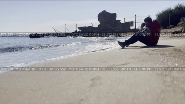 A man with a camera goes to the sandy shore and sits on the sand near the surf. Privacy by the sea. Стоковое видео, видеограф Ярославский Максим / Фотобанк Лори
