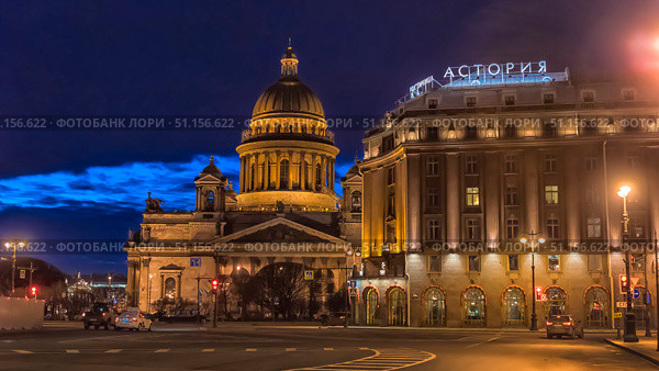 A night photo of St. Petersburg with a view of St. Isaac's Cathedral and the Astoria Hotel. The frame is filled with the atmosphere of an evening city: the majestic dome of the cathedral is illuminated with golden lights, emphasizing the architectural beauty and scale of the building. Nearby is the facade of the historic Astoria Hotel with a bright neon sign reflecting the rich history and status of this place. In the foreground is a deserted street with rare cars and traffic lights, adding dynamics and creating a sense of calm and comfort of the night city. The deep blue color of the sky and the soft lighting of the lanterns create a harmonious combination and emphasize the romantic atmosphere of St. Petersburg. (2020 год). Редакционное фото, фотограф Анна Кривицкая / Фотобанк Лори