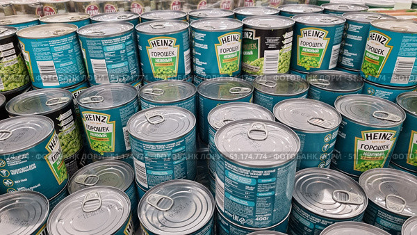 A photograph of supermarket shelves lined with cans of Heinz green peas. The easy-to-open metal cans come in signature green and turquoise packaging with the Heinz logo and the words "Tender Peas." The can lids with pull tabs are clearly visible in the foreground, and the labels with the ingredients and nutritional value of the product are in the background. The photo conveys an atmosphere of abundance and choice in the store, reflecting the theme of trade, consumption, and food packaging. (2021 год). Редакционное фото, фотограф Анна Кривицкая / Фотобанк Лори
