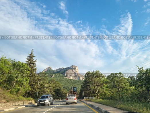 A picturesque section of the highway on the southern coast of Crimea. Ahead, a majestic view of the Kush-Kaya rock massif rises above the green forest. Cars and construction equipment move along the road. On both sides of the highway there is dense vegetation, trees, and tall grass. The sky is clear, with light cirrus clouds emphasizing the scale of the mountains and the vastness of the southern landscape. The scene conveys a sense of freedom, nature, and travel along Crimean routes. (2021 год). Редакционное фото, фотограф Анна Кривицкая / Фотобанк Лори