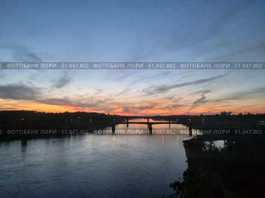 A picturesque view of the river in the evening. On the horizon are the warm shades of the sunset, smoothly transitioning from bright orange and pink to a deep blue sky. Light cirrus clouds stretch across the sky, creating the effect of soft brushstrokes. In the center of the frame you can see the bridge connecting the banks, its dark silhouette clearly stands out against the glowing horizon. On the banks of the river and on the bridge itself, lights are lit, reflecting in the calm water surface. In the right part of the foreground is the dark silhouette of a cliff with vegetation. The atmosphere of the picture conveys the peace and beauty of the sunset hour. Стоковое фото, фотограф Анна Кривицкая / Фотобанк Лори
