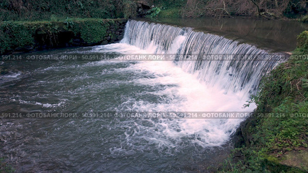 A powerful flow of water cascades over a man-made weir near Cascata do Outeiro. The scene showcases the dynamic interplay of nature and human influence in Oliveira de Azemeis. Стоковое фото, агентство Ingram Publishing / Фотобанк Лори