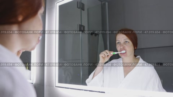 A red-haired woman brushes her teeth before going to bed in the bathroom in front of a large mirror. Стоковое видео, видеограф Арзюков Вадим Николаевич / Фотобанк Лори