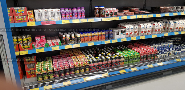 A refrigerated shelf in a supermarket with a wide range of drinks and dairy products. Brightly colored packages of juices, smoothies, milkshakes, yogurts and herbal drinks are arranged in neat rows on the shelves. You can see packages of different colors: green, red, pink, white and black, which creates a colorful palette and emphasizes the diversity of the assortment. On the lower shelves are packages of juices and nectars in cardboard packaging, above - bottles of protein drinks and smoothies. On the right, you can see yogurts in plastic cups and cardboard packaging. Below the shelves are attached price tags with information about the cost of goods. The atmosphere of the photo conveys the everyday look of a grocery store, the orderliness and availability of goods. (2019 год). Редакционное фото, фотограф Анна Кривицкая / Фотобанк Лори
