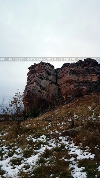 A rock with a characteristic layered structure surrounded by autumn nature. In the foreground, bushes with yellowed leaves are visible, as well as islands of snow on the ground, indicating late autumn or early winter. The rock has a rough texture, many cracks and stratifications, which makes it a unique geological formation.The landscape is typical for the Perm region, where the Ural mountain ranges are combined with forests and open landscapes. Possible location - Vetlan Rock, Chertovo Gorodishche or Stone City. Стоковое фото, фотограф Анна Кривицкая / Фотобанк Лори