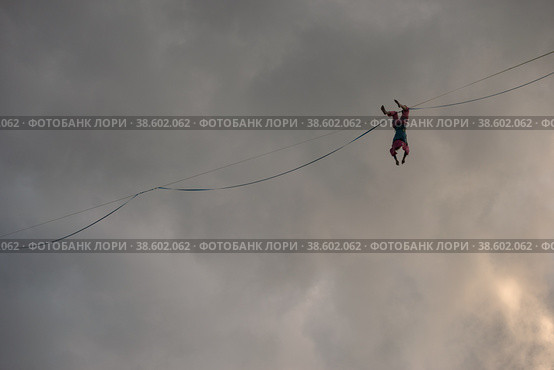 A rope-walker girl hangs upside down on the highline against the background of a dramatic sky. She is resting (2021 год). Редакционное фото, фотограф Happyphotons / Фотобанк Лори