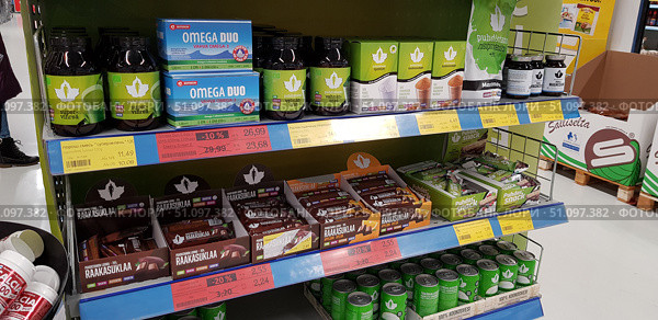 A shop window with products from the Finnish brand Puhdistamo. The shelf is neatly filled with superfoods, dietary supplements and natural products for healthy eating. On the upper shelves you can see jars of green powders Supervihreä (a mix of super greens) and Vehnänoras (wheat grass), Omega Duo capsules with omega-3, as well as boxes of coconut sugar and OptiMSM (methylsulfonylmethane). On the right is a large package of rice protein and jars of magnesium. On the middle level are bars of raw chocolate Raakasuklaa and protein bars Puhdas Snack. Below are tins of coconut water. All products are made in branded green and white packaging, emphasizing the environmental friendliness, naturalness and organic origin of the products. Discounts and promotions are highlighted on the price tags, which creates the atmosphere of an affordable and modern health food store. (2019 год). Редакционное фото, фотограф Анна Кривицкая / Фотобанк Лори