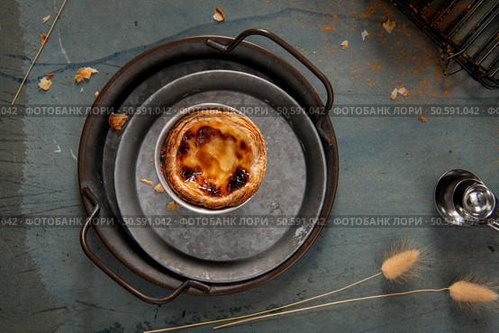 A single Pastel de Nata, resting on a rustic metal tray, with a hint of cinnamon sprinkled on top. Стоковое фото, агентство Ingram Publishing / Фотобанк Лори