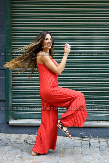 A stylish and confident woman strikes a playful pose in a vibrant red jumpsuit that perfectly showcases her flair and joy, all against a bright green backdrop that adds to the lively scene. A woman in a bright red jumpsuit poses playfully outdoors, radiating joy and confidence. Стоковое фото, агентство Ingram Publishing / Фотобанк Лори