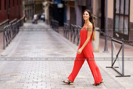 A stylish woman dressed in a vibrant red jumpsuit is elegantly strolling through a picturesque urban alleyway, exuding a remarkable aura of confidence and grace as she enjoys her surroundings. An Elegant Woman Dressed in a Striking Red Suit Walking Gracefully on an Urban Street. Стоковое фото, агентство Ingram Publishing / Фотобанк Лори