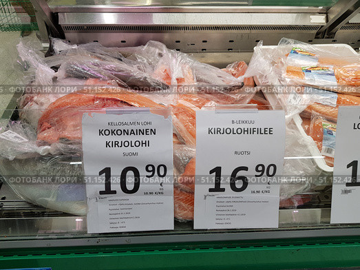 A supermarket counter with chilled packaged fish. In the foreground are large pieces and whole rainbow trout (kirjolohi) wrapped in film. Above the fish are price tags: on the left is the price for a whole fish from Finland - €10.90/kg, on the right is the price for a rainbow trout fillet from Sweden - €16.90/kg. In the background are plastic trays with packaged fillets. The image illustrates the selection of fresh fish in the sales area, the contrast in prices and product cutting options. (2019 год). Редакционное фото, фотограф Анна Кривицкая / Фотобанк Лори
