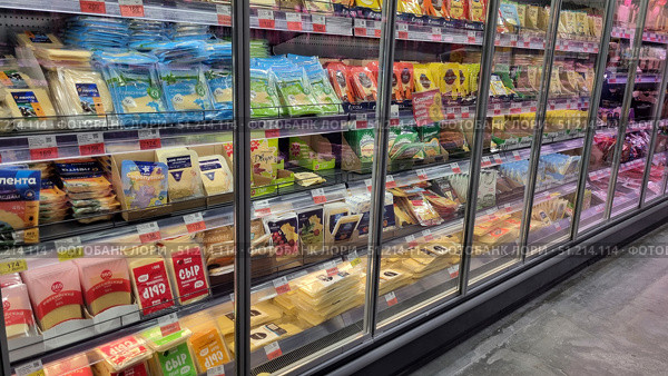 A supermarket display case with a large assortment of sliced ​​and packaged cheeses. The shelves feature various brands and types of cheese: Russian, cream, Gouda, Cheddar, Mozzarella, Edam and others. The packages are brightly colored - blue, green, red, yellow, which makes the display attractive to customers. All products are neatly laid out in a refrigerated section with transparent doors, which emphasizes freshness and proper storage conditions. The price tags indicate promotions and discounts, which creates the feeling of a real store. Редакционное фото, фотограф Анна Кривицкая / Фотобанк Лори