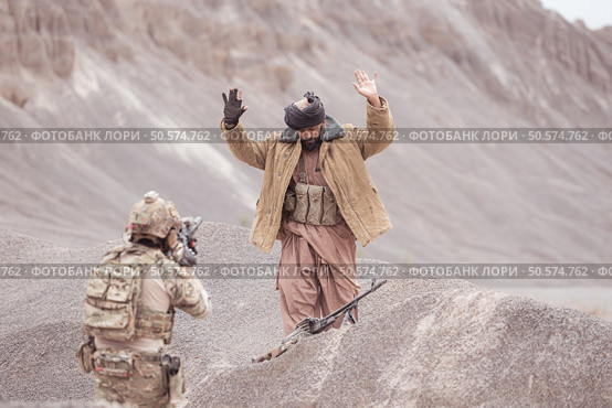 A Taliban soldier stands with a machine gun, Raise hands in surrender to the allied soldiers, In the desert mountain terrain battlefield. Стоковое фото, агентство Ingram Publishing / Фотобанк Лори