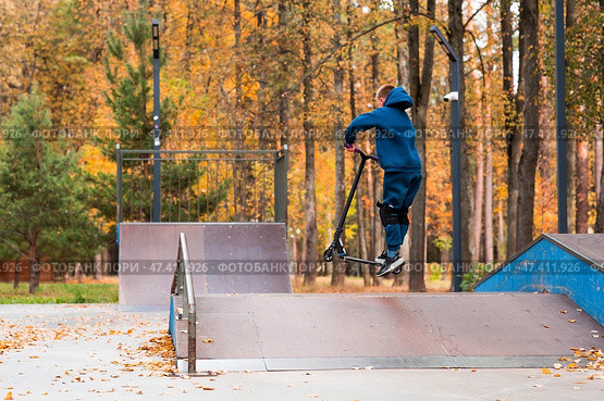 A teenager on a stunt scooter on a playground in a skate park. A child in a tracksuit performs a trick in an autumn park while jumping. Стоковое фото, фотограф Photojuli / Фотобанк Лори