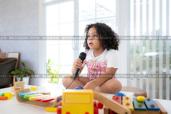 A tiny girl sits on a toy table, clutching a microphone, and sings cheerfully in the living room. Стоковое фото, агентство Ingram Publishing / Фотобанк Лори