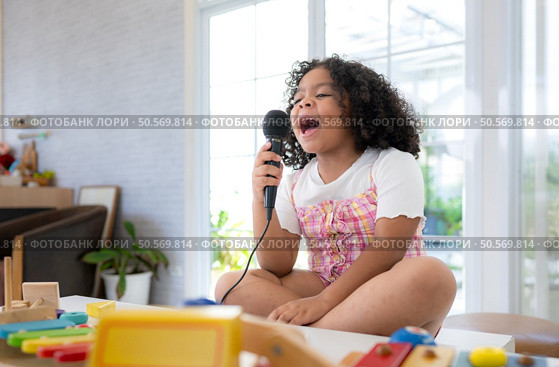 A tiny girl sits on a toy table, clutching a microphone, and sings cheerfully in the living room. Стоковое фото, агентство Ingram Publishing / Фотобанк Лори
