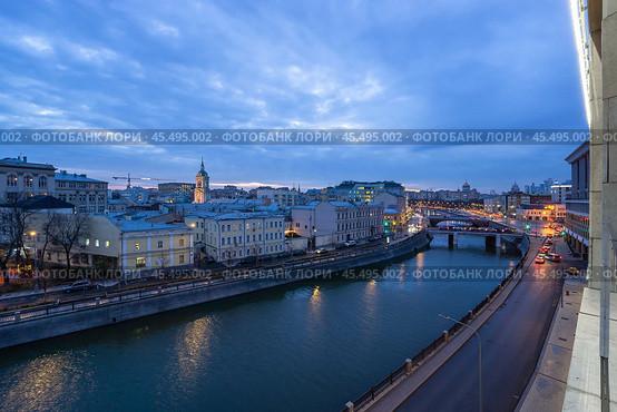 A tranquil city river is highlighted at dusk, reflecting the glowing lights of historic buildings along its banks. A bridge connects both sides, adding charm to the landscape. Стоковое фото, фотограф Алексей Климкин / Фотобанк Лори