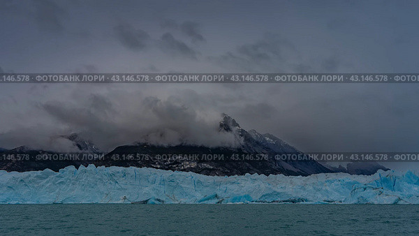 A wall of blue ice with cracks and sharp peaks stretched across the glacial lake. Ripples on the turquoise water. Mountains in clouds and fog. Perito Moreno glacier. El Calafate. Argentina. Стоковое фото, фотограф Вера Ивановна Кубарева / Фотобанк Лори