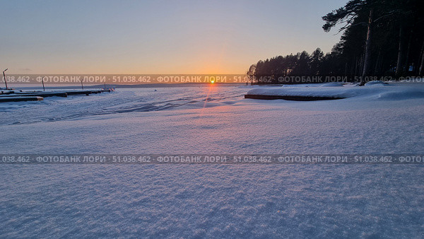 A winter sunset over a frozen lake. The sun is setting on the horizon, painting the snow in soft pink and gold hues. In the foreground is a smooth, shiny blanket of snow, sparkling in the evening light. On the left are the docks extending into the lake, and on the right is the dark silhouette of a pine forest. The atmosphere of peace and quiet, typical of a frosty winter evening. Стоковое фото, фотограф Анна Кривицкая / Фотобанк Лори