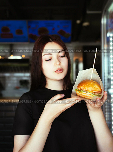 A woman in a black t-shirt looks at a hamburger in her hand with interest (2024 год). Редакционное фото, фотограф Сахарова Мария Юрьевна / Фотобанк Лори
