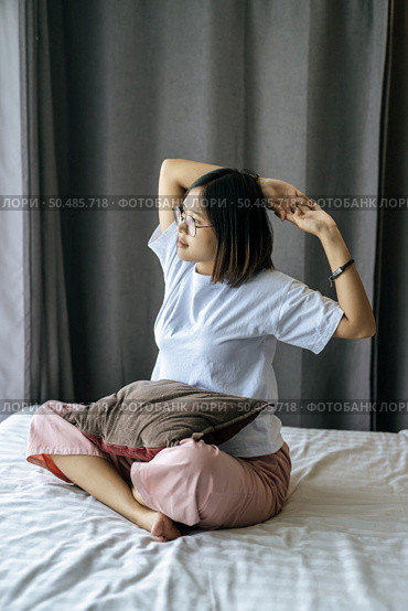 A woman in a white shirt sitting on the bed and raising both arms. Стоковое фото, агентство Ingram Publishing / Фотобанк Лори