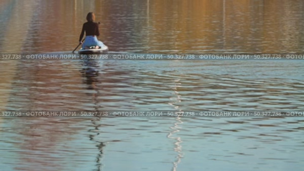A woman in blue jeans and black sweatshirt sits on her knees on a sup board. Back view, she is calmly floating on an inflatable board on a serene lake surrounded by calm water. Стоковое видео, видеограф Воловодов Владимир / Фотобанк Лори