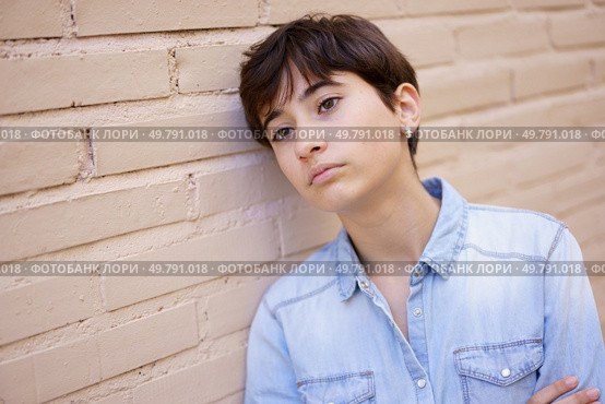 A young individual with stylishly cut short hair is leaning thoughtfully against an appealing beige brick wall, lost in contemplation and reflecting on a myriad of deep emotions within. A Young Person Engaged in Contemplation as They Lean Against a Wall, Deep in Thought. Стоковое фото, агентство Ingram Publishing / Фотобанк Лори